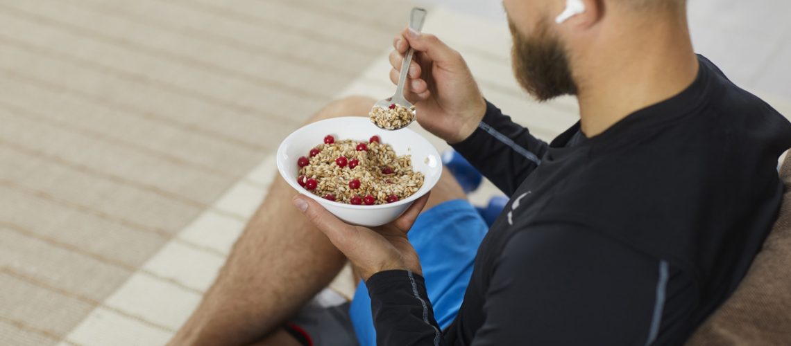 Athlete enjoying healthy meal rich in fiber, protein and vitamins. Fit young man sitting on floor in living room, relaxing after fitness workout, eating natural vegetarian granola, listening to music